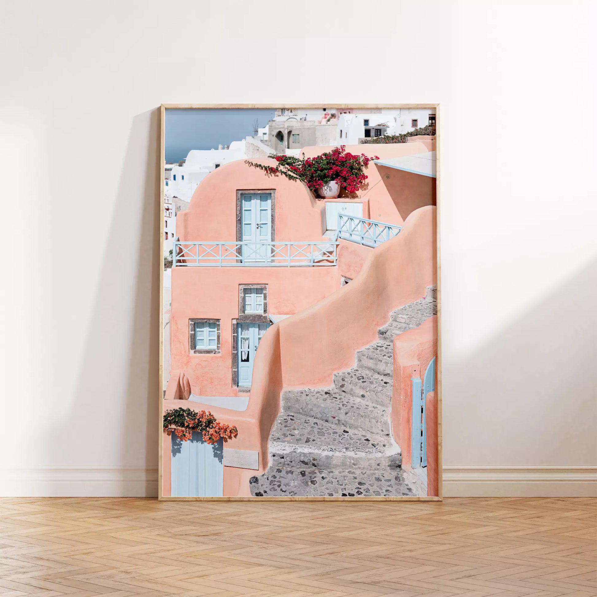 サントリーニ島のパステルピンクの建物と水色のドア、赤いブーゲンビリアが咲く地中海建築アート。白壁の街並みと石畳の小道が見える風景。