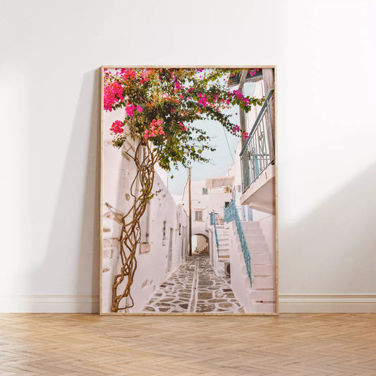 白壁の建物が並ぶ石畳の小道に、鮮やかなピンクのブーゲンビリアが咲き誇り、青い手すりの階段が印象的な地中海風の街並み。伝統的なキクラデス建築と自然が調和した魅力的な景観。