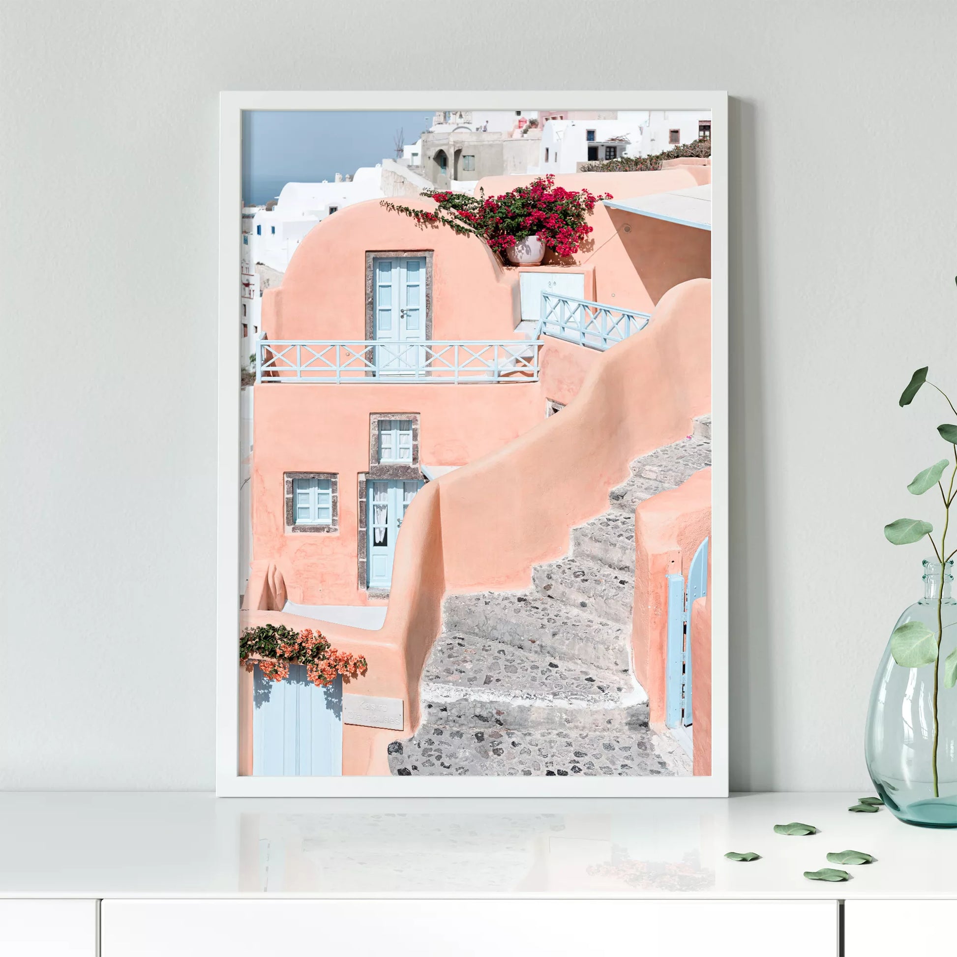 サントリーニ島のパステルピンクの建物と水色のドア、赤いブーゲンビリアが咲く地中海建築アート。白壁の街並みと石畳の小道が見える風景。