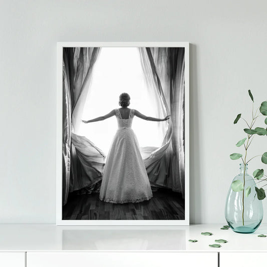 Black and white image of a bride in a lace wedding dress standing at an open window